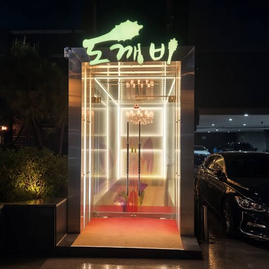 강남 도깨비 쩜오의 입구 전경, 밤에 초록색 도깨비 네온 간판 아래 흰 조명 테두리의 유리문 안으로 샹들리에와 붉은 바닥이 보임